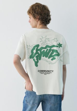 Mladý muž s kučeravými vlasmi, ktorý nosí biele oversize tričko so zeleným abstraktným dizajnom a nápisom "COMMUNITY", v kombinácii s modrými džínsami.