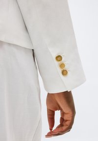 Χέρι που ακουμπά δίπλα σε μανίκι λευκού σακακιού με τρία χρυσά κουμπιά στην μανσέτα, ορατό τμήμα ταιριαστού λευκού παντελονιού.