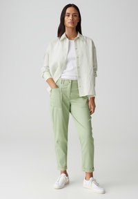 Pantalon droit vert clair avec poches avant, associé à une chemise texturée blanc cassé, portée par-dessus un top blanc et des baskets blanches.