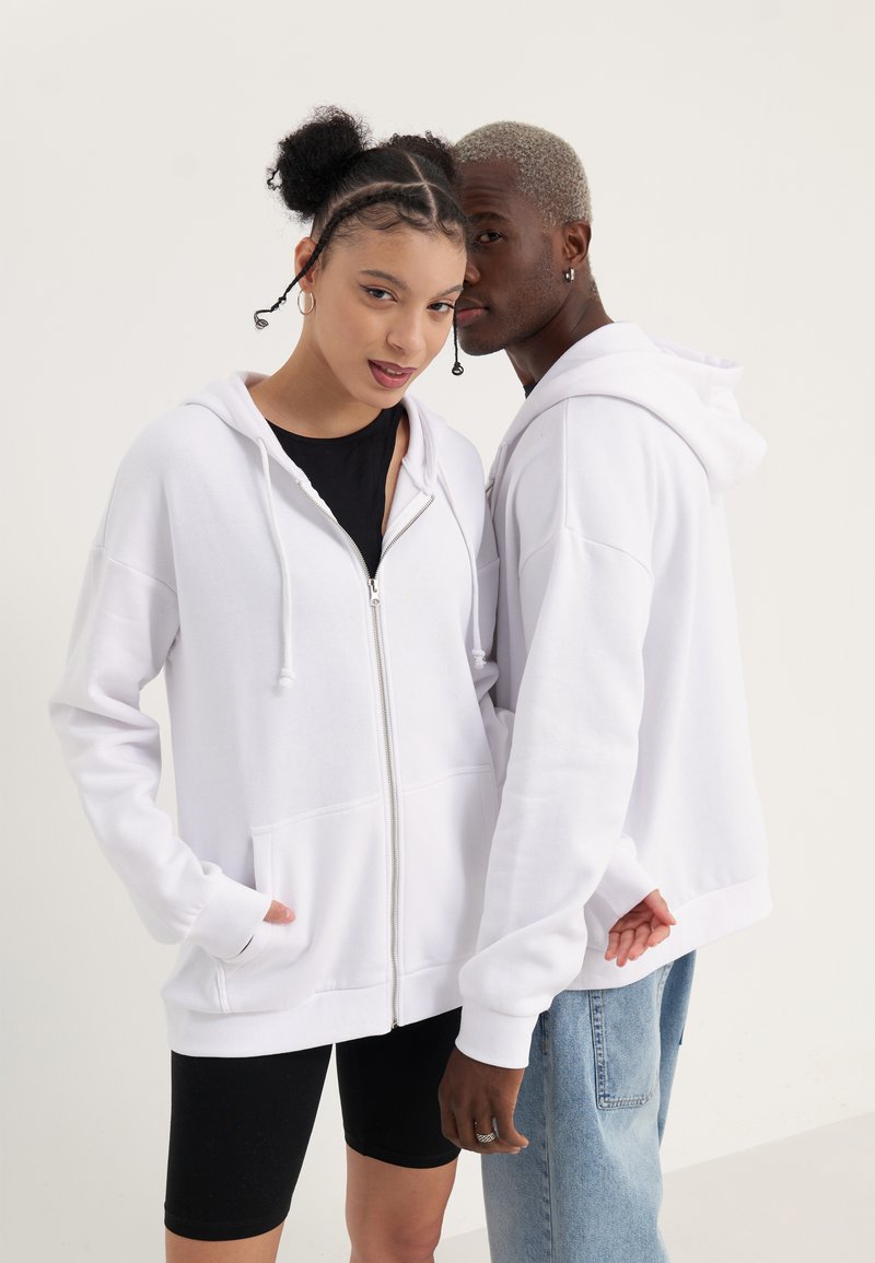 Felpa bianca con zip e cappuccio regolabile, progettata con un fit rilassato e tasche frontali, indossata sopra un top nero e abbinata a pantaloni corti neri.
