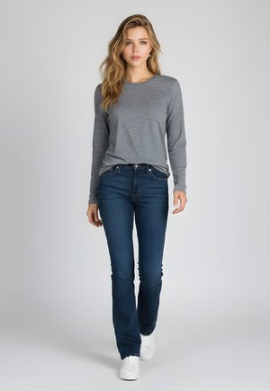 Mujer joven con cabello largo y rubio que lleva una camiseta de manga larga gris, vaqueros azul oscuro y zapatillas blancas, caminando hacia adelante sobre un fondo liso.