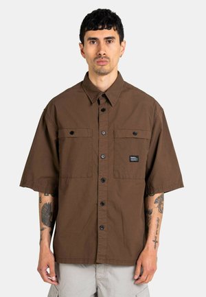 Man with short dark hair wearing a loose brown short-sleeve button-up shirt and light gray pants, showing tattoos on both forearms.