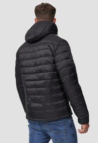 Doudoune noire avec capuche, présentant des panneaux matelassés et une finition lisse. Le design comprend des poignets élastiques et une coupe décontractée.