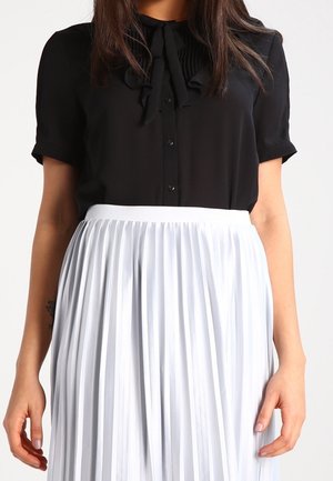 Woman wearing a black short-sleeve blouse with front buttons and a bow tie paired with a high-waisted white pleated skirt.