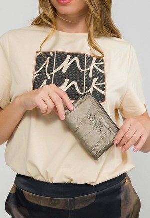 Donna che tiene in mano un portafoglio metallico argentato con cerniera e logo "Anekke", indossa una t-shirt beige con testo grafico bianco e nero e pantaloni a fantasia.