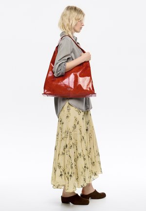 Czerwona, błyszcząca skórzana torba tote z szerokim otwarciem i wydłużonym kształtem, noszona na ramieniu, na tle jasnej, kwiatowej spódnicy i szarego topu.