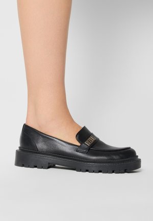 Foot wearing a black leather loafer with thick lug sole and gold logo detail on strap against a plain gray background.