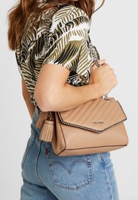 Nainen, jolla on päällään vihreä-valkoinen kuviollinen paita ja siniset farkut, pitää käsissään vaaleanruskeaa tilkkutäkkilaukkua, jossa on hopeinen ketjuhihna ja tupsuyksityiskohta.