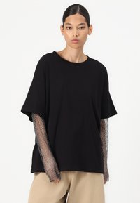 Černé oversized tričko s krátkými rukávy, s černými mesh rukávy jako akcent. Měkká textura se uvolněným střihem, spárované s béžovými kalhotami.