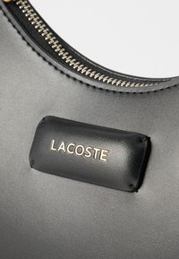 Fekete bőr táskafül rész ezüst cipzárral és varrott fekete folttal, amelyen arany betűkkel a „LACOSTE” márkanév olvasható.