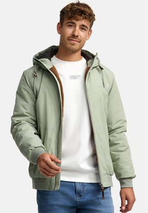 Cazadora bomber verde claro con capucha, que presenta un cierre de cremallera en la parte delantera, puños elásticos y sutiles acentos marrones. Llevada sobre una camiseta blanca.