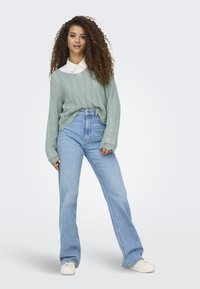 Maglione lavorato a maglia verde chiaro con una camicia bianca con colletto sotto, abbinato a pantaloni in denim blu a vita alta con gambale svasato e sneaker bianche.