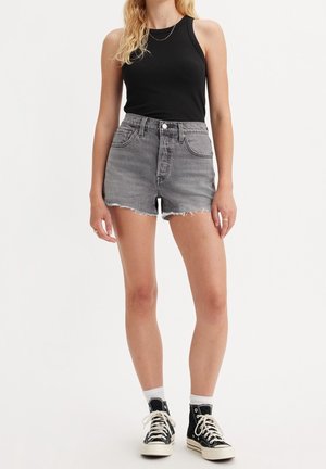 Kvinde iført sort ærmeløs top, grå frynset denimshorts, hvide sokker og sorte høje sneakers, står foran en ensfarvet baggrund.