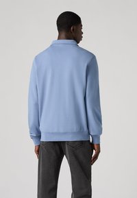 Himmelblauer Polo-Sweatshirt mit Kragen, langen Ärmeln und gerippten Bündchen, kombiniert mit dunkelgrauen Jeans. Glatte Textur.