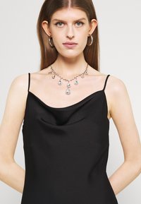 Camisola de cetim preta com decote em U, alças finas e detalhe drapeado. Usada com um colar de cristais em gota e brincos de argola.