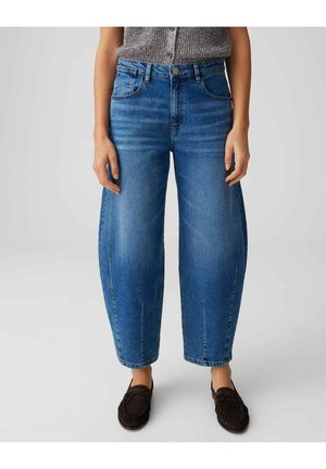 Persoon draagt cropped wijde blauwe jeans en donkere mocassins, staand tegen een effen lichte achtergrond.