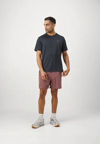 Camiseta desportiva de manga curta cinza escuro com textura canelada, combinada com calções leves cor-de-rosa. Pés calçados com ténis cinzentos, a posar sobre um fundo claro.