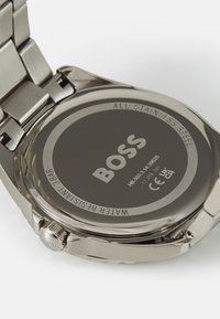 Montre en acier inoxydable avec un fond de boîtier rond gris mat. Gravée avec "BOSS" et des détails de résistance à l'eau. Bracelet en maillons métalliques.