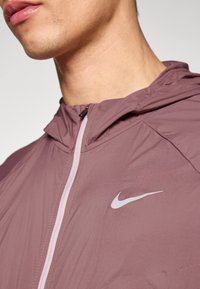 Nike'i kapuutsiga jope summutatud lavendlivärvitoonis, iseloomulik kerge materjal, tõmblukk ja kontrastne heledam õmblus. Rinnal ikooniline logo.