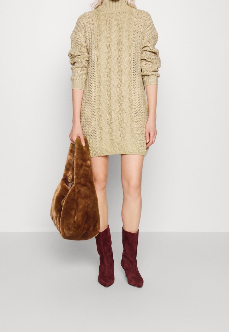 Personne portant une robe pull en maille torsadée beige et des bottines bordeaux, tenant à son côté un grand sac en fausse fourrure marron.
