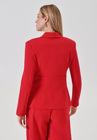 Rode op maat gemaakte blazer met een gestructureerde pasvorm, voorzien van een gesneden kraag, lange mouwen en een tailleband. Soepele stoftextuur en strakke lijnen.