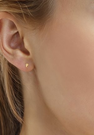 Close-up of ear wearing small gold stud earring shaped like letter "P" with light brown hair tucked behind ear.