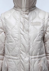 Světlé béžové prošívané vatované sako s prošitým vzorem, nastavitelnou kapucí, dvěma předními kapsami a logovou nášivkou na levé horní straně.