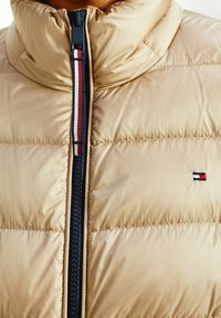Beige vadderad jacka med hög krage. Har en mörk dragkedja och ett randigt accent i rött, vitt och blått. Litet broderat märke på bröstet.