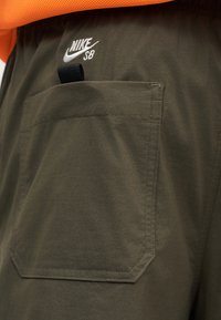 Κεντημένο λογότυπο Nike SB πάνω από μια μεγάλη τσέπη ελιάς με μαύρη υφασμάτινη θηλιά σε παντελόνια ελιάς και πορτοκαλί ένδυμα από πάνω.