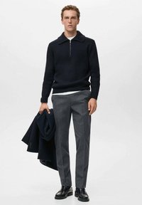 Maglione lavorato a maglia blu navy con colletto a zip, pantaloni grigi a righe e scarpe nere in pelle. Un cappotto blu navy è appoggiato su un braccio.