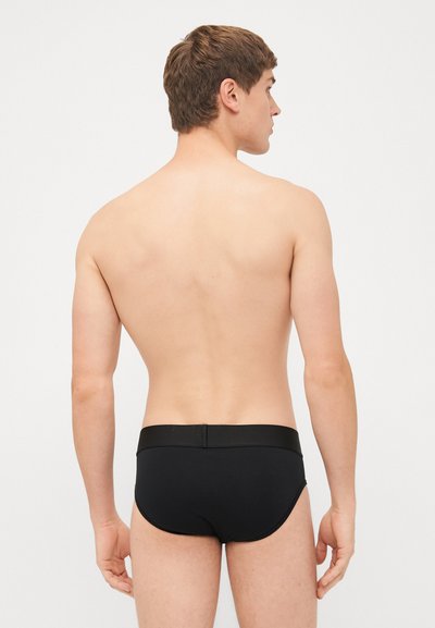 Sorte svømmebukser med en glat tekstur, elastisk talje og minimalistisk design, sett fra baksiden mot en enkel bakgrunn.