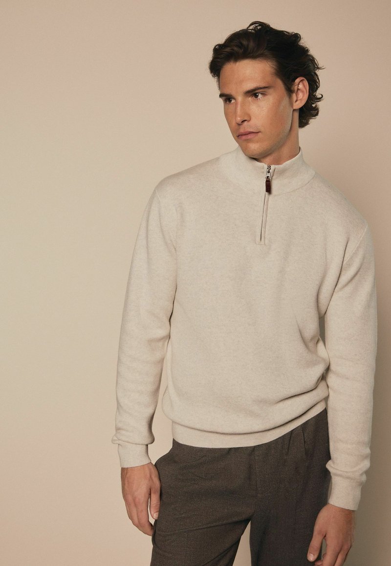 Maglione beige con zip alta e polsini a costine, realizzato in un morbido tessuto a maglia. Caratterizzato da una piccola cerniera con un tirante a contrasto.