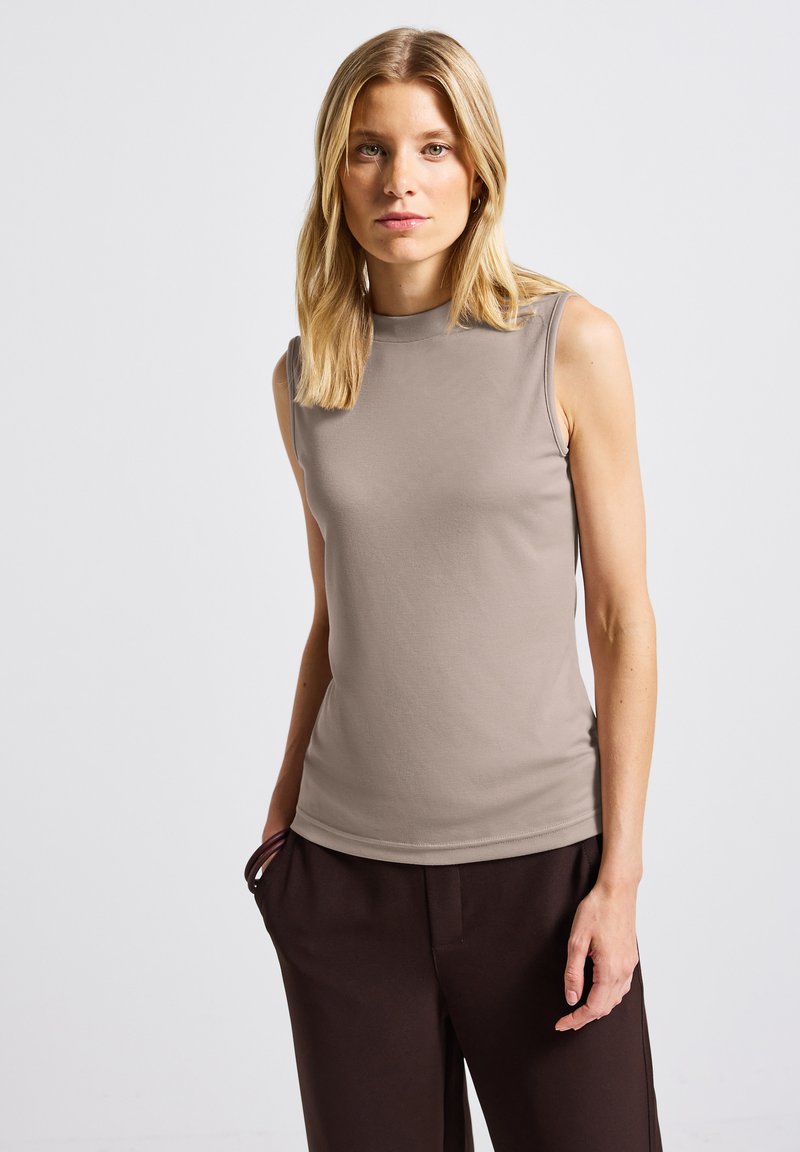 Blond kvinna i ärmlös taupe-top och mörkbruna byxor med händerna i fickorna mot en enkel ljus bakgrund.