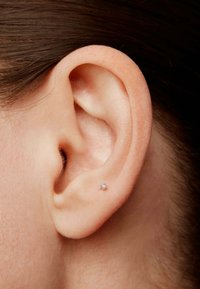 Pequeño pendiente de plata con una gema redonda y transparente, colocado en el lóbulo de una oreja humana. El tono de piel es claro y se pueden ver pelos finos.