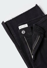 Jeans de mezclilla negra con cremallera, botón de metal y dobladillo deshilachado. Presentan una etiqueta de marca con detalles de tamaño y costuras cuidadas en toda su extensión.