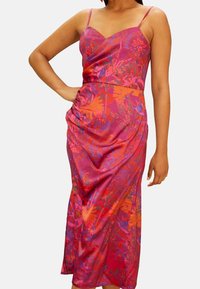 Nainen pukeutuneena hihattomaan, kukalliseen kietaisumekkoon, jossa on ohuet hihnat ja värisävyinä vaaleanpunainen, punainen, oranssi ja violetti, käsi lanteilla, seisomassa yksinkertaisen taustan edessä.