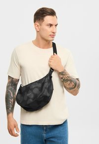 Geantă de umăr neagră cu un model texturat, închidere cu fermoar și curea ajustabilă. Fabricată dintr-un material durabil, având un logo de marcă proeminent.