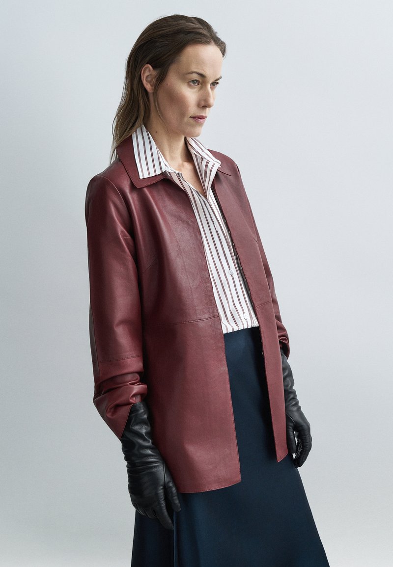 Γυναίκα με μπορντό δερμάτινο μπουφάν, μαύρα δερμ�άτινα γάντια, ριγέ λευκό πουκάμισο και σκουρό μπλε φούστα, στέκεται μπροστά σε έναν απλό ανοιχτόχρωμο φόντο.