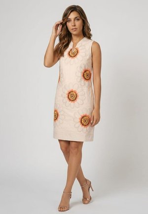 Mujer con vestido beige sin mangas con grandes patrones de flores naranjas y amarillas y sandalias de tacón marrón claro, de pie con una mano cerca del cabello.