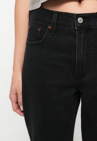 Pantalones vaqueros de mezclilla negra con cintura alta, que cuentan con un bolsillo frontal, herrajes de cobre y detalles de costura sutiles a lo largo de los bordes.