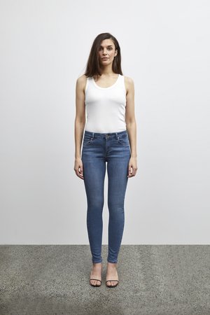 Kvinde med langt brunt hår iført en hvid ærmeløs top, blå stramme jeans og sorte åbne sandaler, der står på en tekstureret gulv.