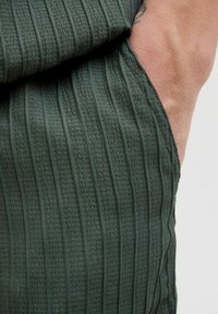 Tissu vert texturé avec des raies verticales et un motif à carreaux subtil, montrant un gros plan des détails de la manche d'un vêtement.