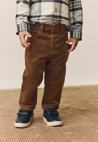 Pantaloni in corduroy marrone con una superficie testurizzata, abbinati a una camicia a quadri con bottoni. Sneakers nere con strap in velcro completano l'outfit.