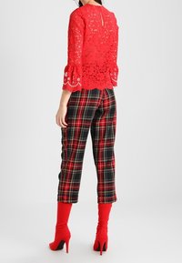 Top in pizzo rosso con maniche a tre quarti, orlo scallop, abbinato a pantaloni a quadri neri e rossi. L'outfit presenta stivali con tacco alto rossi.