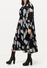 Langes schwarzes Kleid mit weißen und bunten abstrakten Mustern, ausgestattet mit Falten und langen Ärmeln. Kombiniert mit schwarzen Stiefeln mit Absatz.