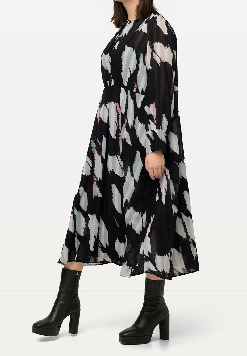 Langes schwarzes Kleid mit weißen und bunten abstrakten Mustern, ausgestattet mit Falten und langen Ärmeln. Kombiniert mit schwarzen Stiefeln mit Absatz.