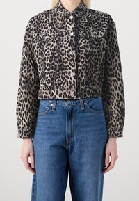 En modell bär en kort leopardmönstrad denimjacka över högmidjade blå jeans, med händerna synliga vid sidorna. Neutral bakgrund.