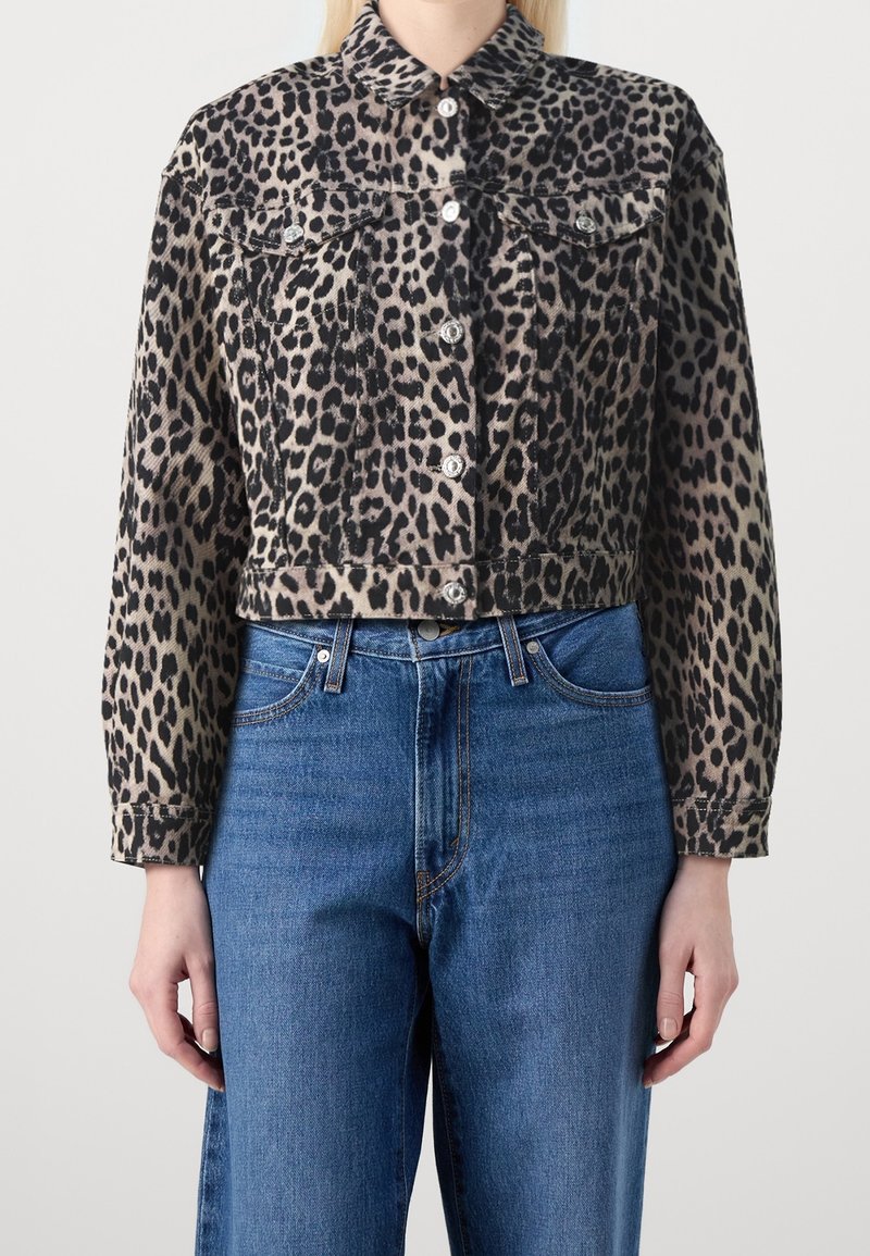 En modell bär en kort leopardmönstrad denimjacka över högmidjade blå jeans, med händerna synliga vid sidorna. Neutral bakgrund.