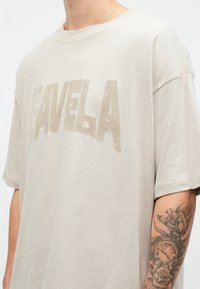 Tricou oversized de culoare bej deschis cu un imprimeu distorsionat "FAVELA" în tonuri de bej mai închis. Mâneci scurte, guler rotund, textură moale din bumbac.