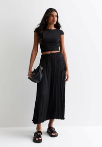 Top negro corto combinado con una fluida falda larga plisada negra. Accesorios incluyen un pequeño bolso negro y sandalias de deslizamiento negras.